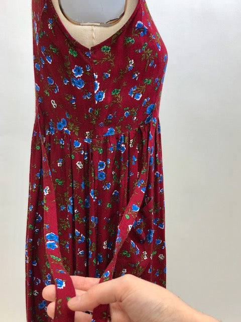 Burgundy sleeveless prairie dress with little blue and white flowers, gathered high doll waist and patch pockets to hold your freshly-picked flowers. Loose fit with double ties at waist to adjust for contouring. Midi length. This adorable dress looks homemade. 