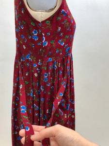 Burgundy sleeveless prairie dress with little blue and white flowers, gathered high doll waist and patch pockets to hold your freshly-picked flowers. Loose fit with double ties at waist to adjust for contouring. Midi length. This adorable dress looks homemade. 