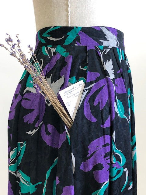 Stunning vintage midi flared skirt with waist pleats, side pockets and center back zipper. Black with purple and green floral print. Gorgeous silky viscose fabric with a sheen pattern. Perfect for dancing, twirling, and loving. Daniel Richie label. 